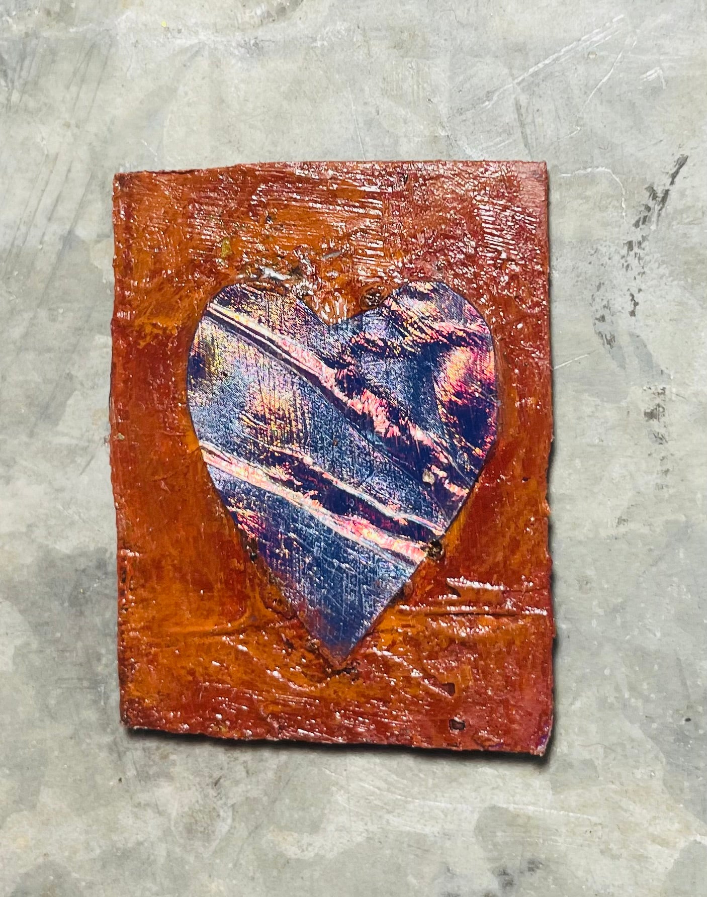 A rectangular magnet with a foil heart shape in the center, attached to a acrylic, reddish-brown background.